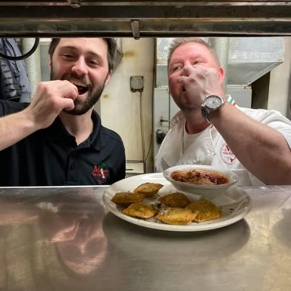 Two chefs in a kitchen, one smiling, with a plate of fried ravioli and marinara sauce.