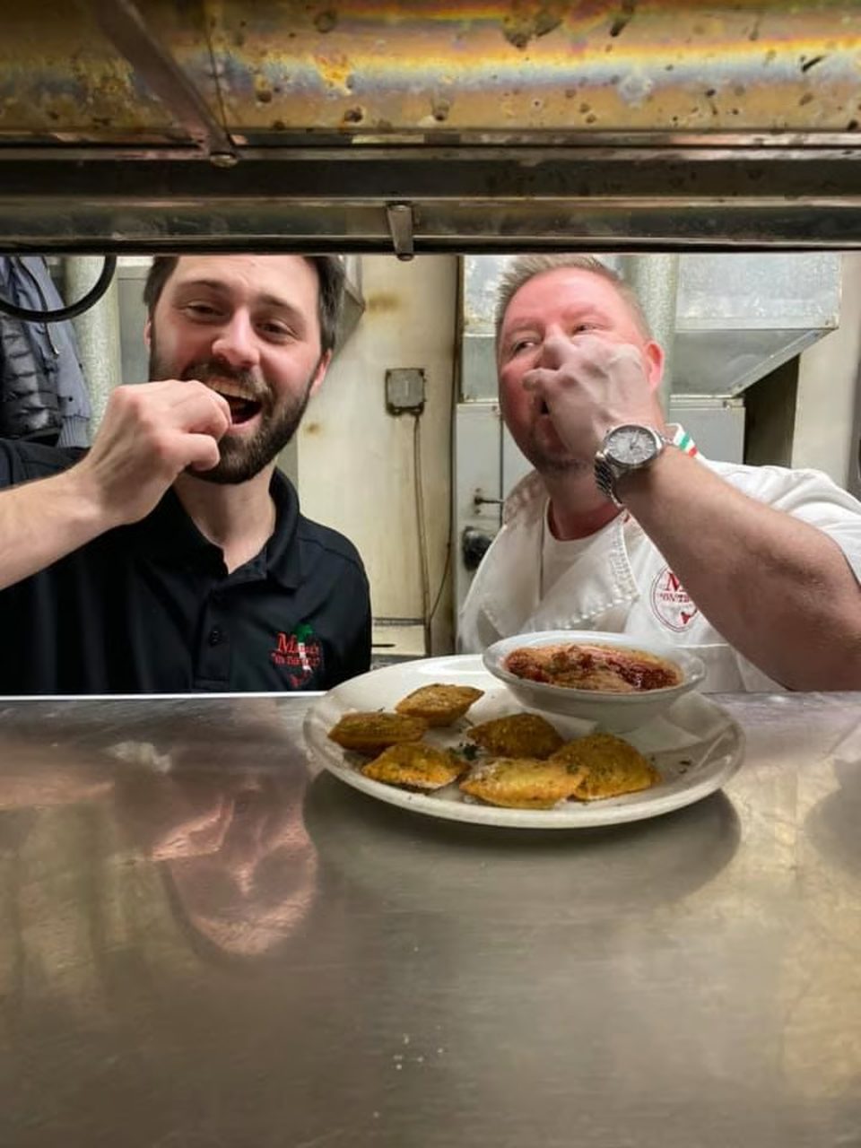 Two chefs in a kitchen, one smiling, with a plate of fried ravioli and marinara sauce.