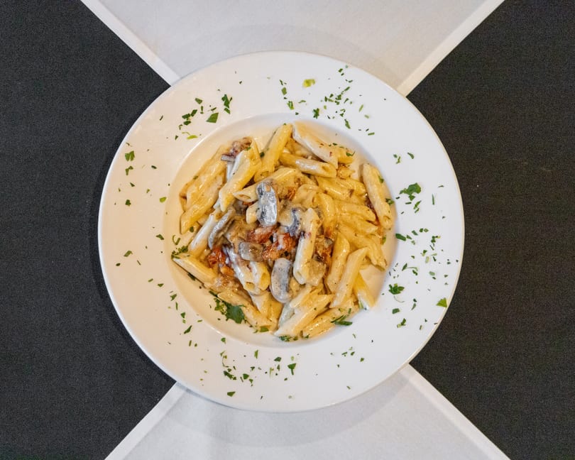 White plate of creamy penne pasta with mushrooms, bacon, and fresh parsley.