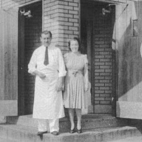Black and white photo of a man in an apron and a woman in a dress standing outside a brick building.