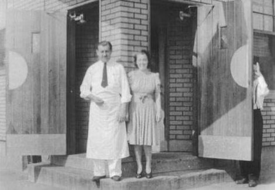 Black and white photo of a man in an apron and a woman in a dress standing outside a brick building.