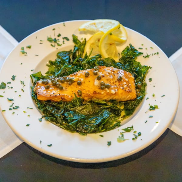 Grilled salmon fillet with capers and lemon slices on a bed of sautéed greens, garnished with parsley.