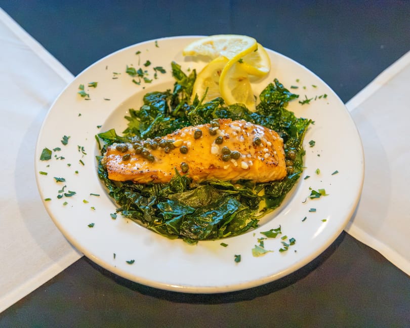 Grilled salmon fillet with capers and lemon slices on a bed of sautéed greens, garnished with parsley.