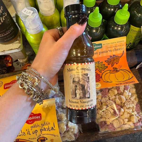 A hand holds a bottle of Mama Campisi's Balsamic Vinaigrette amidst various pasta products.