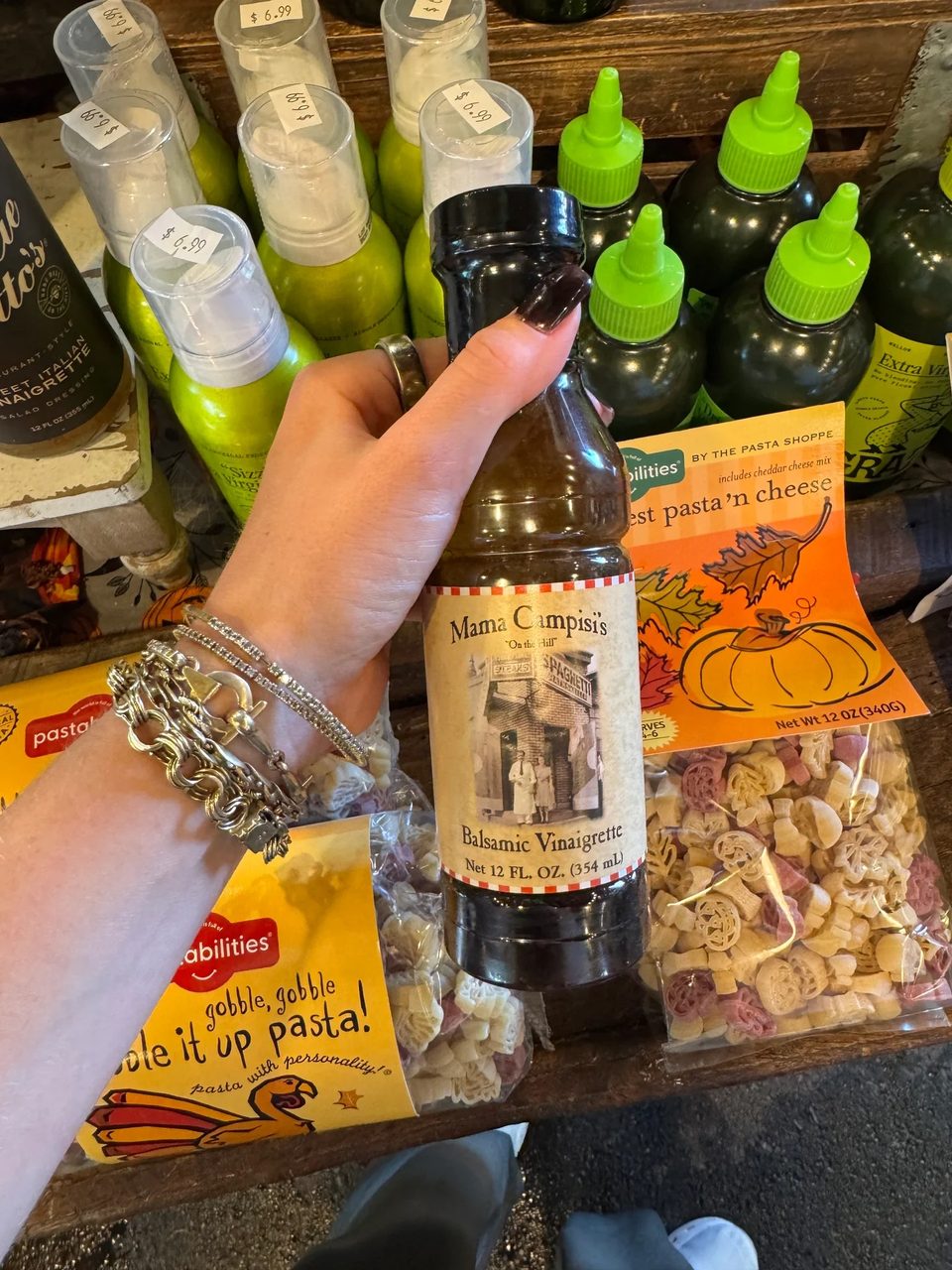 A hand holds a bottle of Mama Campisi's Balsamic Vinaigrette amidst various pasta products.