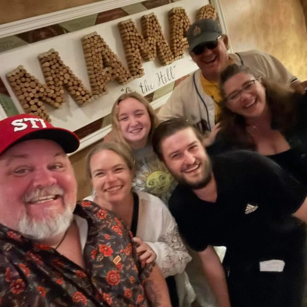Six smiling people pose for a selfie in a restaurant with a "MAMA" sign in the background.