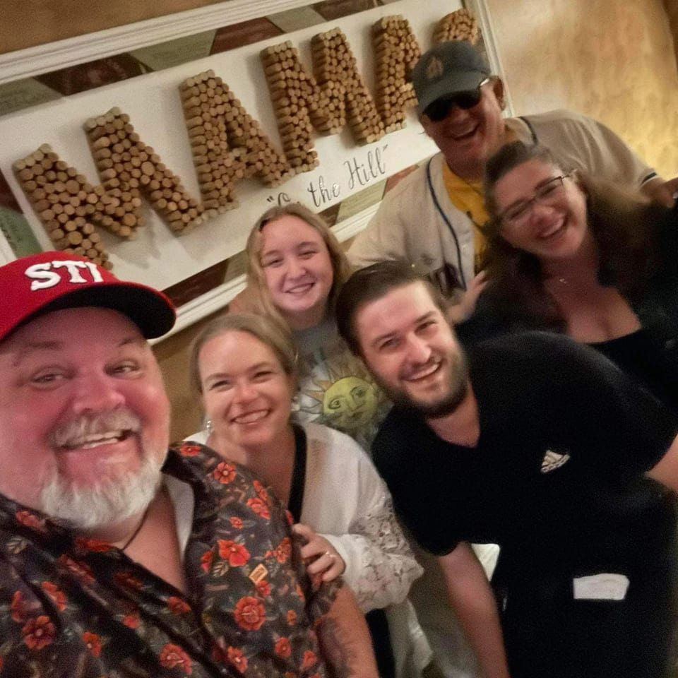 Six smiling people pose for a selfie in a restaurant with a "MAMA" sign in the background.
