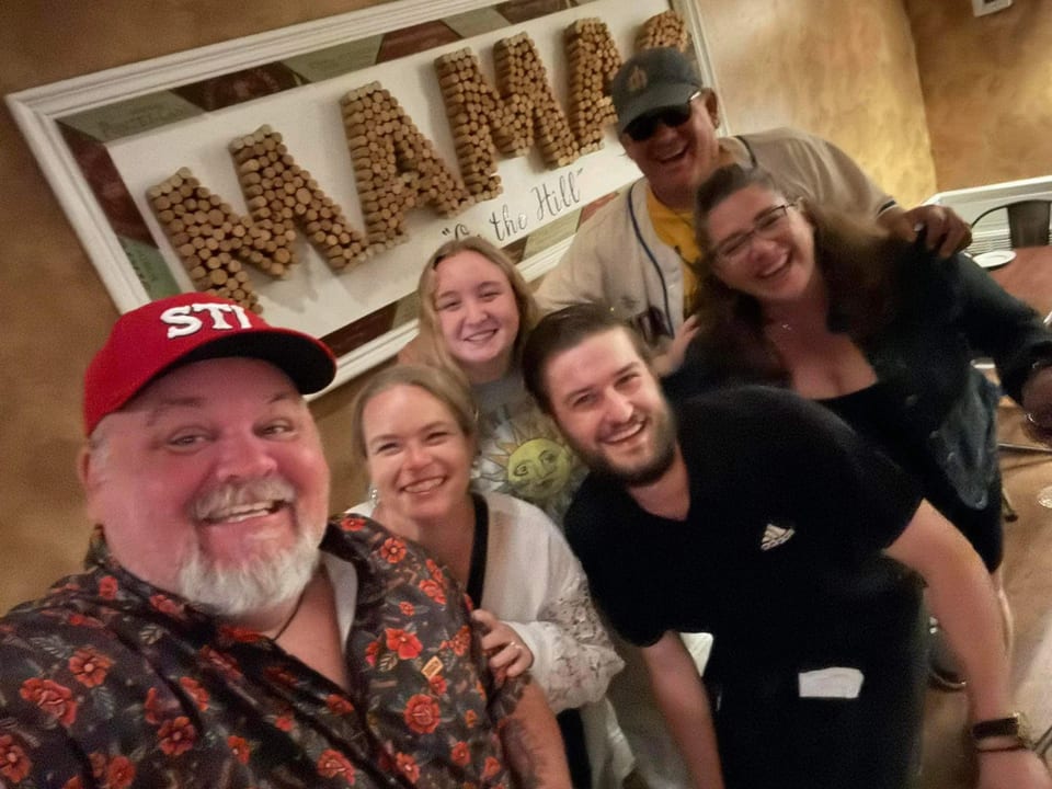 Six smiling people pose for a selfie in a restaurant with a "MAMA" sign in the background.