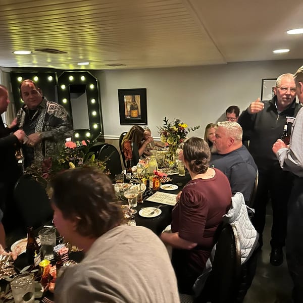 Guests gather around a long table with drinks and floral centerpieces at a lively restaurant event.
