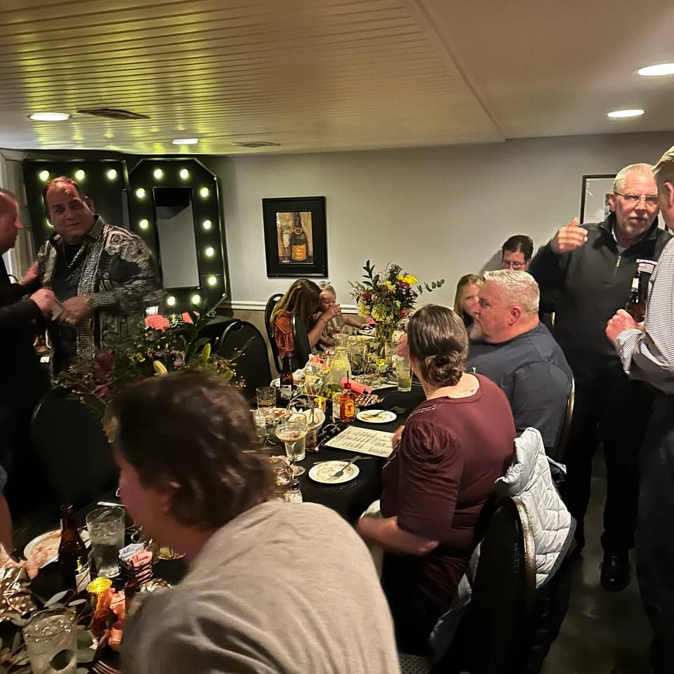 Guests gather around a long table with drinks and floral centerpieces at a lively restaurant event.