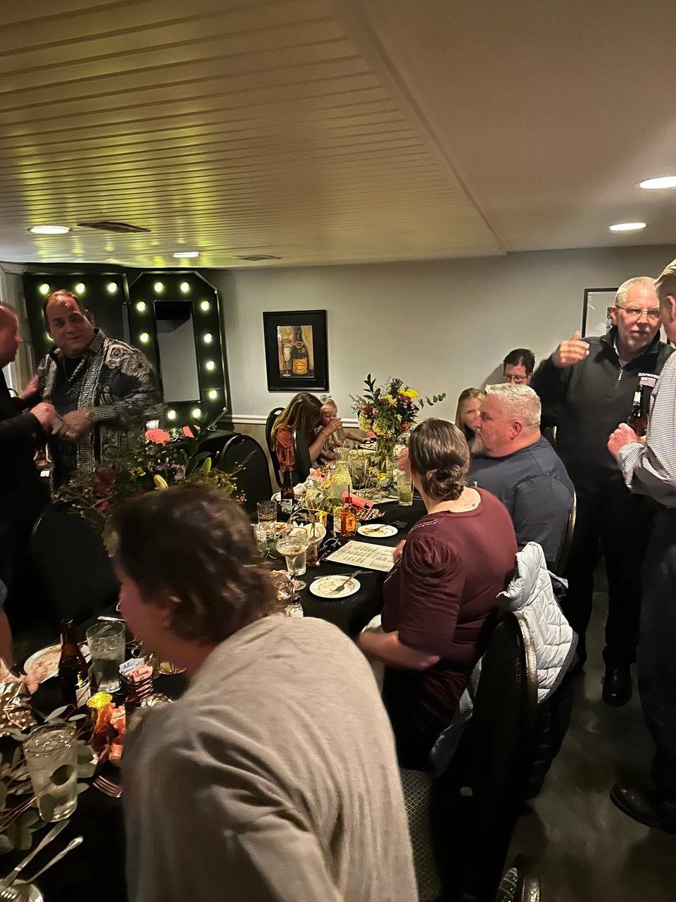 Guests gather around a long table with drinks and floral centerpieces at a lively restaurant event.