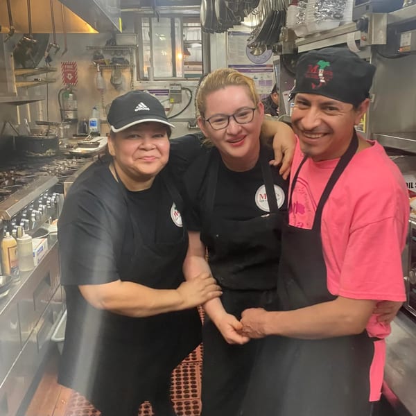 Three smiling kitchen staff members, two women and one man, posing together in a busy restaurant kitchen.
