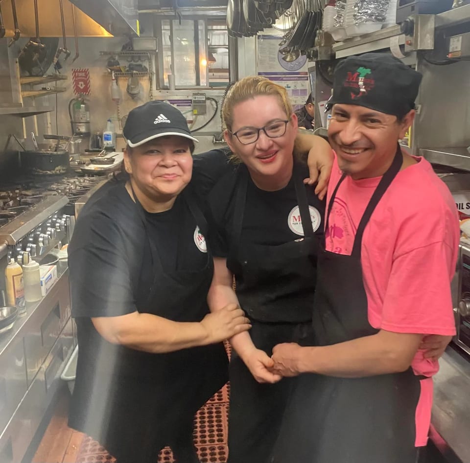 Three smiling kitchen staff members, two women and one man, posing together in a busy restaurant kitchen.