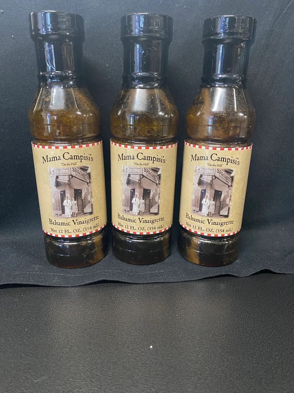 Three bottles of Mama Campisi's Balsamic Vinaigrette with vintage labels on a black surface.
