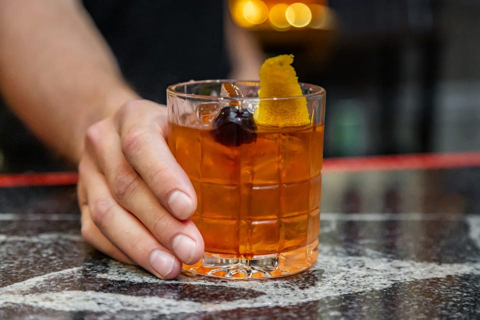 A hand reaching for an Old Fashioned cocktail garnished with a cherry and orange peel on a bar.