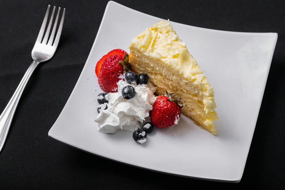 A slice of white chocolate cheesecake with whipped cream, strawberries, and blueberries on a white plate.