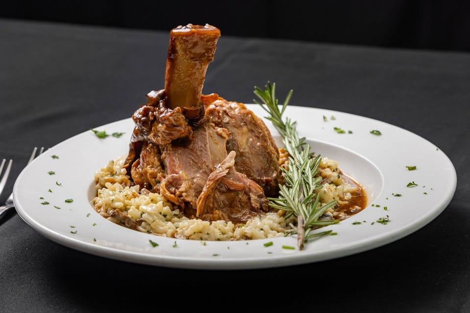 Braised lamb shank with mushroom risotto and rosemary sprigs on a white plate.
