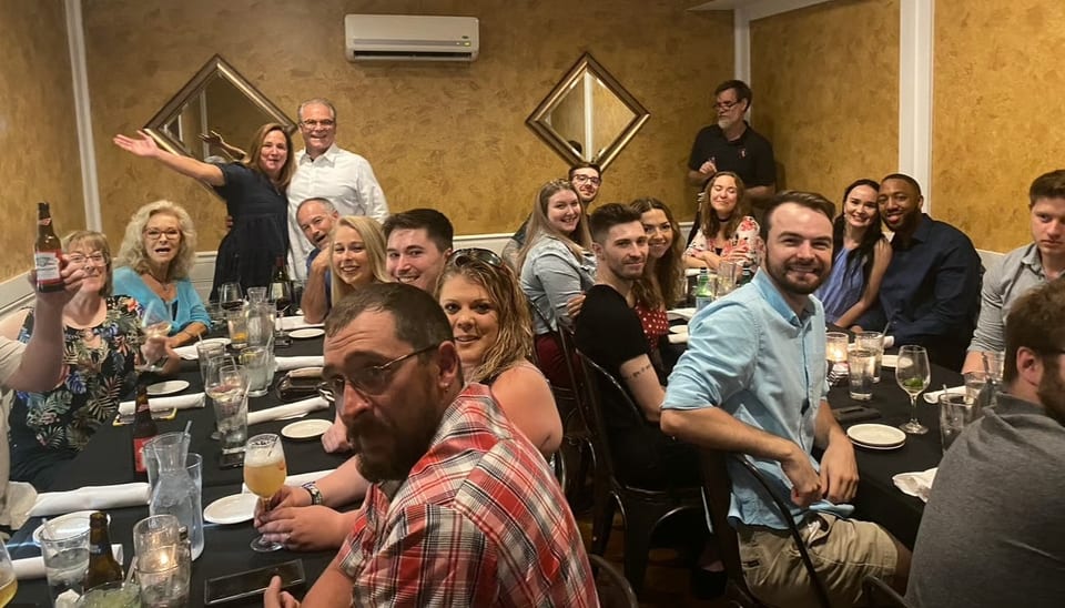 A large group of smiling people gathered around a long table in a restaurant, enjoying a meal.