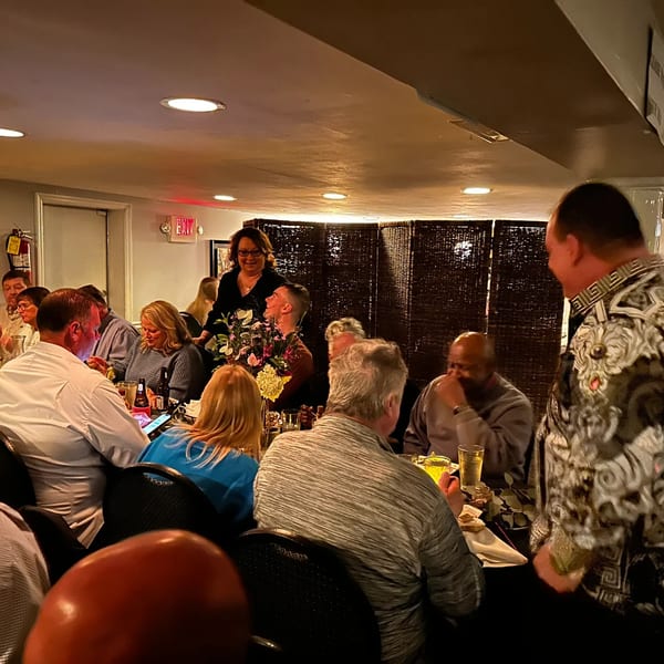 Diners seated at tables in a dimly lit restaurant, with a woman standing and smiling.