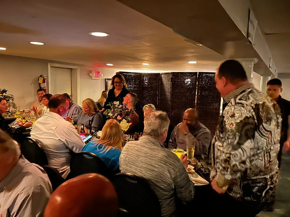 Diners seated at tables in a dimly lit restaurant, with a woman standing and smiling.